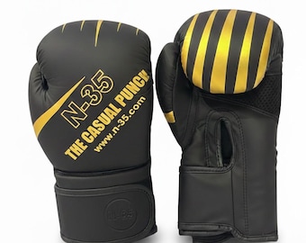 Puede incluir: Un par de guantes de boxeo negros con detalles dorados. El guante izquierdo presenta el texto N-35, THE CASUAL PUNCH y www.n-35.com en letras doradas. El guante derecho muestra rayas doradas verticales en la parte superior y un panel de malla.