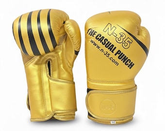 Puede incluir: Un par de guantes de boxeo dorados metalizados con rayas negras en los nudillos. Un guante presenta el texto negro N-35, THE CASUAL PUNCH y www.n-35.com. Las muñequeras incluyen un logotipo circular N-35 y una pequeña etiqueta blanca.