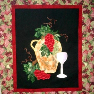 May include: A quilted wall hanging with a black background features a brown ceramic pitcher with a bunch of red grapes and a clear glass of wine. The pitcher and grapes are surrounded by green vines. The background is framed with a red border and a floral pattern.