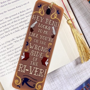 May include: A brown bookmark with a gold tassel. The bookmark features a cartoon illustration of two pistols and the text "Hey Beni! Looks to me like you're on the wrong side of the river!"