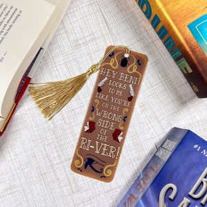 May include: A brown bookmark with a gold tassel. The bookmark features a quote from the book "The Way of Kings" by Brandon Sanderson: "Hey Beni! Looks to me like you're on the wrong side of the river!"