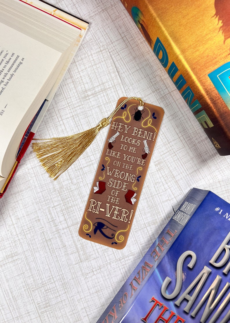 May include: A brown bookmark with a gold tassel. The bookmark features a quote from the book "The Way of Kings" by Brandon Sanderson: "Hey Beni! Looks to me like you're on the wrong side of the river!"