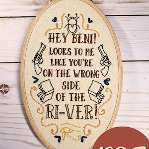 May include: Embroidered hoop art with a beige background and brown text that reads "HEY BENI! LOOKS TO ME LIKE YOU'RE ON THE WRONG SIDE OF THE RI-VER!" with a beetle, guns, and books in the design.