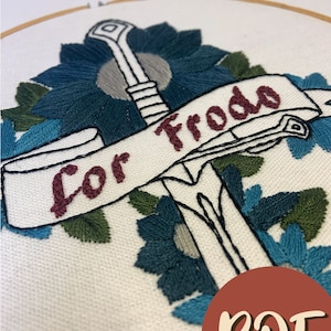 May include: Embroidered hoop art featuring a sword with a banner that reads "For Frodo". The sword is surrounded by blue and green flowers.