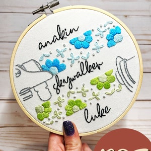 May include: Embroidered hoop art with the text "anakin skywalker luke" in black cursive. The hoop features a stylized hand holding a lightsaber with blue and green flowers surrounding the text.