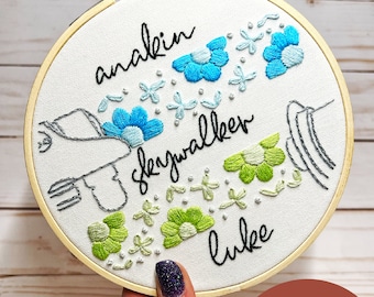 Può includere: Arte di ricamo a cerchio con il testo "anakin skywalker luke" in corsivo nero. Il cerchio presenta una mano stilizzata che tiene una spada laser con fiori blu e verdi che circondano il testo.