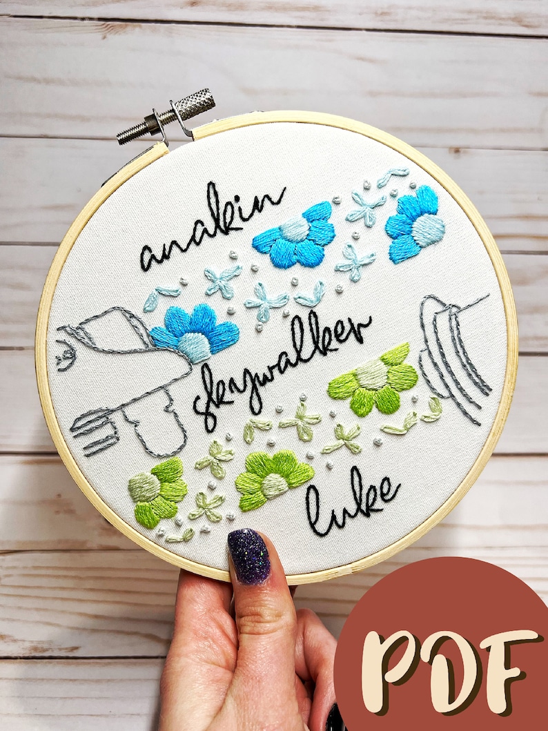 May include: Embroidered hoop art with the text "anakin skywalker luke" in black cursive. The hoop features a stylized hand holding a lightsaber with blue and green flowers surrounding the text.