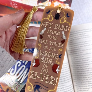 May include: A brown bookmark with a gold tassel. The bookmark features a cartoon beetle, two pistols, and the text "Hey Beini! Looks to me like you're on the wrong side of the ri-ver!"