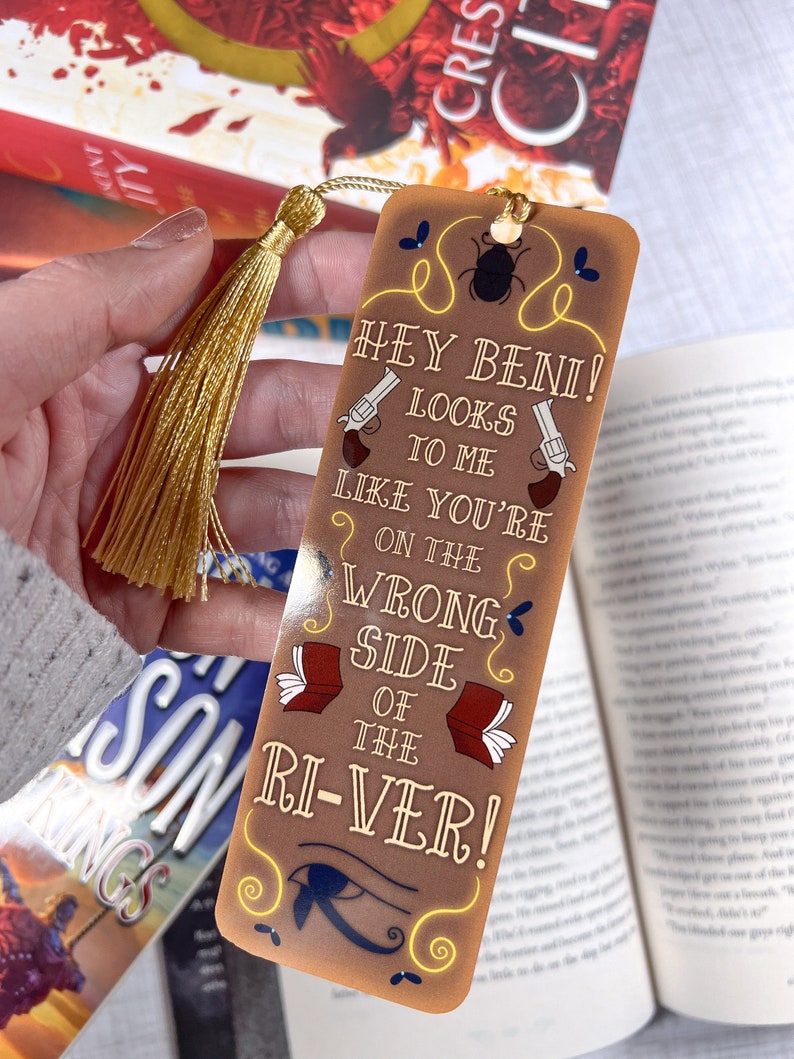 May include: A brown bookmark with a gold tassel. The bookmark features a cartoon beetle, two pistols, and the text "Hey Beini! Looks to me like you're on the wrong side of the ri-ver!"