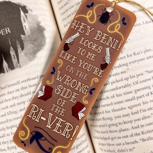 May include: A brown bookmark with a yellow border and a gold tassel. The bookmark features a cartoon illustration of two pistols, a beetle, and a book. The text on the bookmark reads "Hey Beni! Looks to me like you're on the wrong side of the river!"