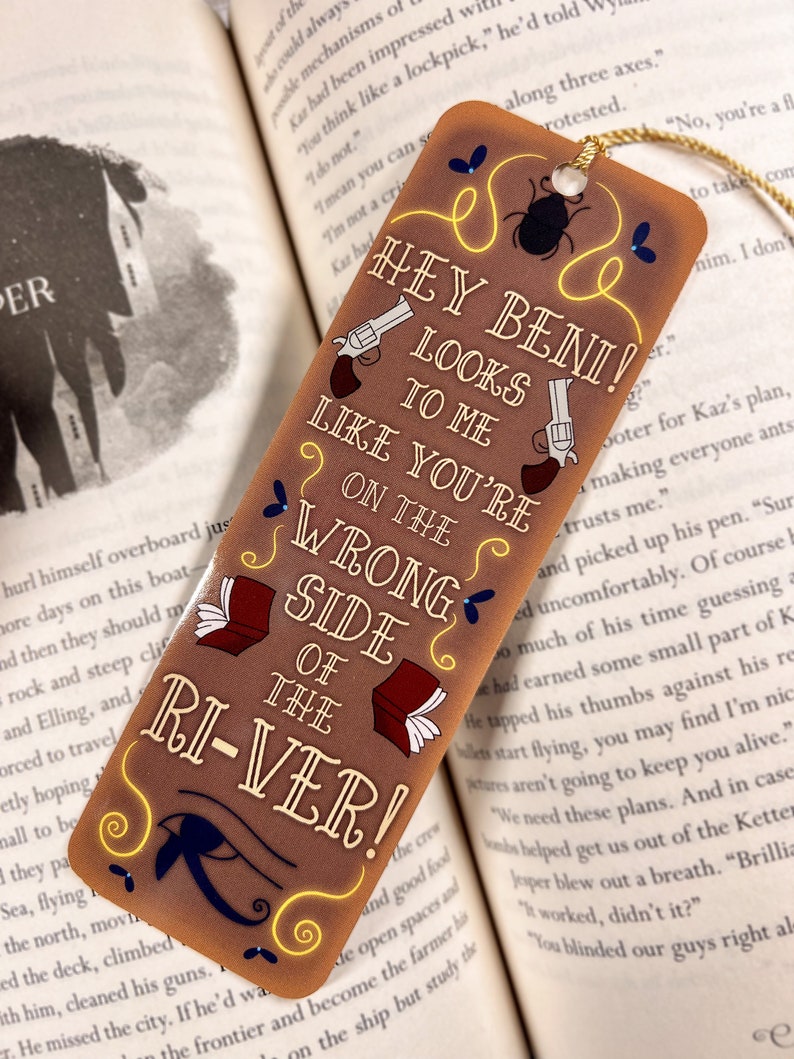 May include: A brown bookmark with a yellow border and a gold tassel. The bookmark features a cartoon illustration of two pistols, a beetle, and a book. The text on the bookmark reads "Hey Beni! Looks to me like you're on the wrong side of the river!"