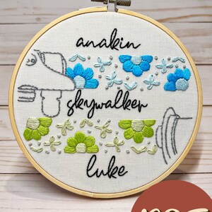 May include: Embroidered hoop art with the text "anakin skywalker luke" in black cursive. The hoop features a stylized drawing of a spaceship with blue and green flowers surrounding it.