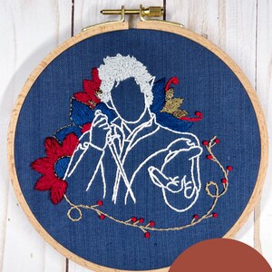 Op de afbeelding: Borduurwerk in een hoepel met een witte omtrek van een persoon met een microfoon, omringd door rode, blauwe en gouden bloemmotieven op een donkerblauwe stoffen achtergrond.