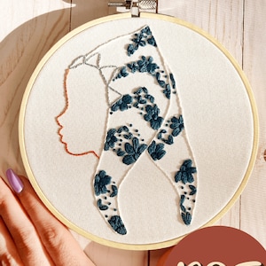 May include: Embroidered hoop art featuring a profile of a person wearing a headscarf with a floral pattern. The headscarf is teal blue and the background is white.