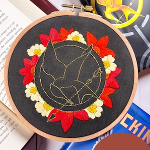 May include: Embroidered hoop art featuring a gold mockingjay with a golden arrow, surrounded by a wreath of red and white flowers. The hoop is on a wooden surface with a book titled "The Final Book of Mockingjay" visible in the background.