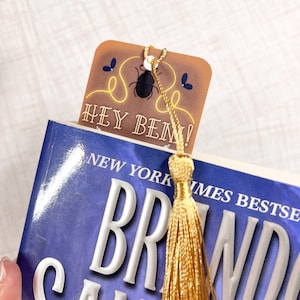 May include: A brown bookmark with a gold tassel. The bookmark features a black beetle illustration and the text "Hey Ben!" in white. The bookmark is on top of a blue book with the text "New York Times Bestseller" and "Brandon Sanderson" on the cover.