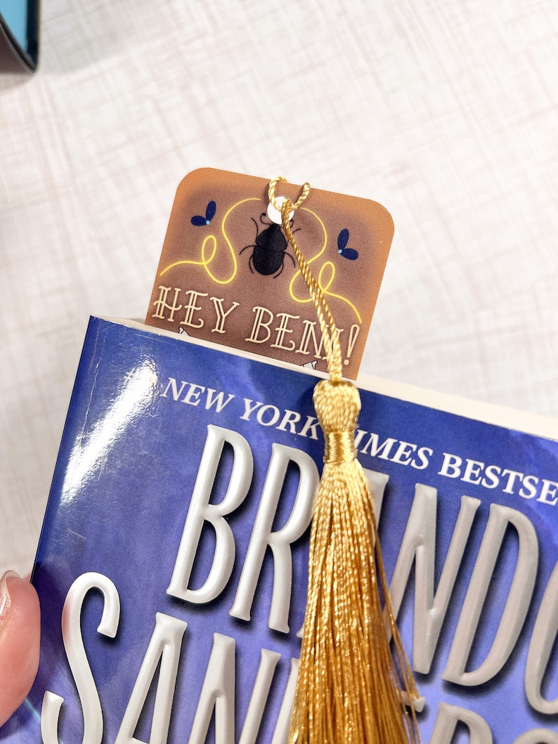 May include: A brown bookmark with a gold tassel. The bookmark features a black beetle illustration and the text "Hey Ben!" in white. The bookmark is on top of a blue book with the text "New York Times Bestseller" and "Brandon Sanderson" on the cover.