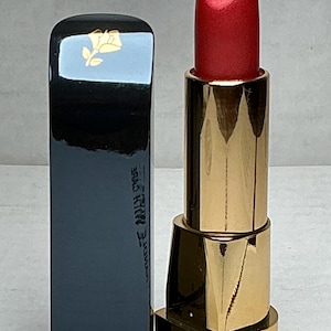May include: A red lipstick with a gold-colored base and a black rectangular cap. The cap has a gold rose emblem and the brand name "Lancôme" is printed on the bottom. The lipstick is open, revealing the red color.