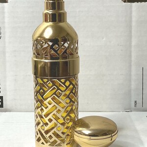 May include: A gold-colored glass bottle with a spray nozzle and a decorative, openwork design. The bottle is sitting on a white surface next to a gold-colored cap.
