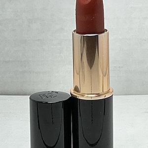 May include: Open lipstick tube with a dark red lipstick bullet. The lipstick has a gold-colored metal base and a black cap. The cap is off to the side, revealing the lipstick. The background is a light gray.