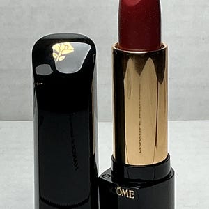 May include: A red lipstick with a black cap and gold-colored tube. The lipstick is open, revealing the deep red color of the product. The cap is glossy black with a gold emblem. The brand name is visible on the base.