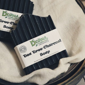 Op de afbeelding: Een zwarte zeep in blok met een wit label waarop staat "DEVANA ORGANICS Tea Tree-Charcoal Soap NO ARTIFICIAL COLOURS OR FRAGRANCES". De zeep is handgemaakt en heeft een geribbelde textuur.