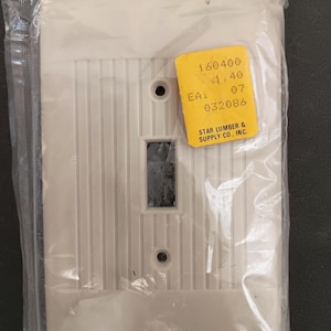 Pode incluir: Uma nova tampa de interruptor de luz branco sujo com ranhuras verticais, embalada em plástico transparente. Um autocolante amarelo com o texto "160400", "1.40", "EA: 07", "032086" e "STAR LUMBER & SUPPLY CO., INC." está anexado.
