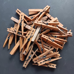 Puede incluir: Un montón de pinzas de ropa de madera con muelles metálicos. Las pinzas son de varios tonos de marrón y beige, sobre un fondo gris oscuro. Las pinzas están dispuestas de forma aleatoria.