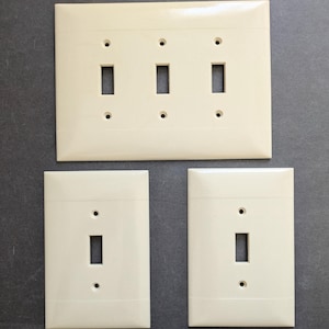 May include: A set of three off-white light switch wall plates. One large rectangular plate features three vertical toggle switch openings and six screw holes. Two smaller rectangular plates each feature one toggle switch opening and two screw holes.