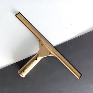 May include: A gold-coloured window squeegee with a black rubber blade. The handle has a rounded end and is inscribed with text. The squeegee is angled across a white and grey surface.