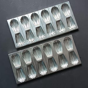 May include: Two rectangular, silver madeleine baking trays. Each tray has 12 shell-shaped moulds. The trays are made of metal and are designed for baking the classic French madeleine biscuits. The trays are on a dark surface.