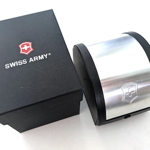 May include: A black rectangular Swiss Army branded box with a red and white shield logo, positioned next to a curved black and silver display case featuring a metallic silver center panel with an embossed Swiss Army shield emblem on the front.