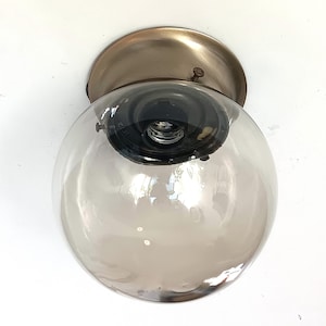 Op de afbeelding: Een plafondlamp in vintage-stijl met een heldere, bolvormige glazen kap met een rookgrijze tint. Het heeft een messingkleurige metalen basis en een zichtbare gloeilamp. De armatuur is gemonteerd op een wit oppervlak. De glazen bol heeft een diameter van ongeveer 20 cm.