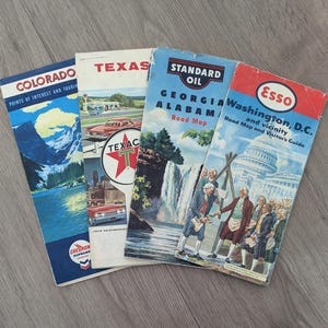 May include: Four vintage paper road maps featuring Colorado, Texas, Georgia-Alabama, and Washington, D.C. The covers display colorful illustrations of mountain landscapes, classic cars, waterfalls, and historical figures in front of the U.S. Capitol building.