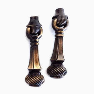 Puede incluir: Dos tiradores de armario antiguos de color bronce con un diseño decorativo. Cada tirador presenta un asa curvada, un centro acanalado y una base con diseño en espiral. Los tiradores se muestran sobre un fondo blanco.