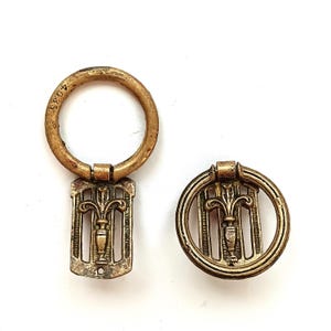 May include: Two antique brass key fobs. One has a rectangular design with a fleur-de-lis motif and a ring. The other is a circular design with the same motif. The ring has the number "4035" engraved on it.