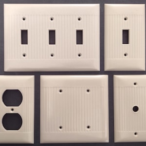 Puede incluir: Un conjunto de cinco placas de pared de color blanco hueso con una textura acanalada vertical. El set incluye una placa para interruptor triple, una para interruptor simple, una para enchufe doble, una placa ciega y una para puerto de cable redondo.