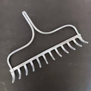May include: A silver-colored garden rake with a curved handle and a row of tines. The rake is made of metal and appears to be used for gardening or landscaping tasks. The tines are evenly spaced and slightly curved.