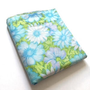 May include: Folded floral bed sheet with a pattern of large blue and white daisies with light blue and lavender accents on a green background. The sheet is made of a soft fabric, likely cotton, and is neatly folded.