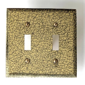 May include: A square double light switch wall plate featuring a metallic gold finish with a dark, intricate crackle texture. The plate includes two rectangular cutouts for toggle switches and four matching gold screws for installation.