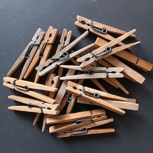 May include: A collection of wooden clothespins with metal springs. The clothespins are various shades of brown, from light tan to darker wood tones. The metal springs are dark gray. The clothespins are arranged in a pile.