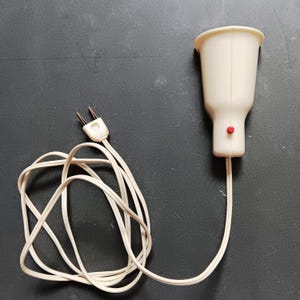 Puede incluir: Un electrodoméstico eléctrico vintage de color crema con una parte superior acampanada y un botón rojo. El aparato está conectado a un cable blanco y a un enchufe de dos clavijas. El cable está enrollado.