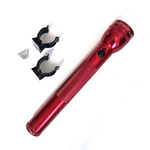 Op de afbeelding: Een rode Mag-Lite zaklamp met een zwarte aan/uit-knop, twee zwarte montageclips en diverse zilveren schroeven. De zaklamp is ongeveer 30 cm lang.