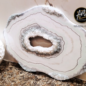 Könnte beinhalten: Weiße Achat-Geoden-Wandkunst mit silbernem Glitzerfinish und goldener Umrandung. Die Geode hat eine große Öffnung in der Mitte. Die Kunst ist mit "Art Territory" signiert und enthält den Social-Media-Handle "@art_territory_charlotte".