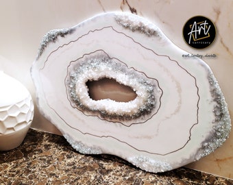 May include: White agate geode wall art with a silver glitter finish and a gold outline. The geode has a large opening in the center. The art is signed "Art Territory" and includes the social media handle "@art_territory_charlotte".