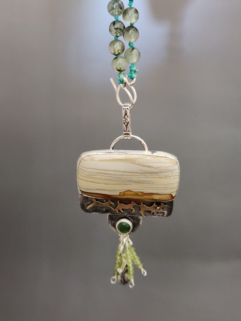 May include: A silver pendant necklace with a rectangular white and brown agate stone, a green stone, and a silver tassel. The necklace is made of green and white beads.