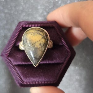 May include: A silver ring with a teardrop-shaped stone. The stone has yellow, grey, and black hues. The ring is set in a silver band and is presented in a purple velvet ring box.