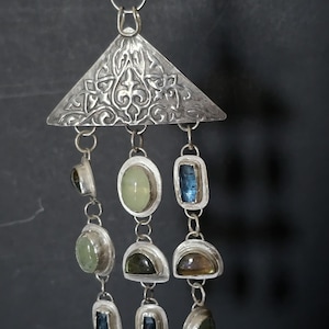 May include: A silver pendant with a triangular top featuring ornate carvings. Below, various gemstones in oval, rectangular, and half-moon shapes dangle from delicate chains. The gemstones are in shades of green, blue, and brown.