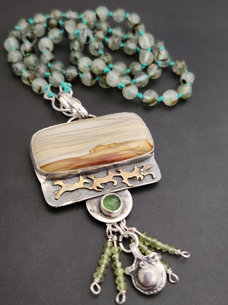 May include: A silver pendant necklace with a rectangular agate stone, green gemstone accents, and a silver charm with a beaded tassel. The necklace is made with green and black beads.