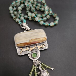 May include: A silver and gold necklace with a large, rectangular, brown and white agate pendant. The pendant is surrounded by a silver frame with gold accents. The necklace is made of green beads and has a silver and gold charm with a green stone.